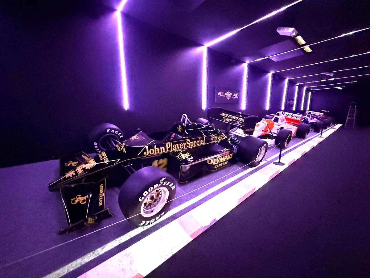 Exposição de carros de Fórmula 1 em um ambiente com iluminação roxa, destacando um carro preto com patrocínio John Player Special.