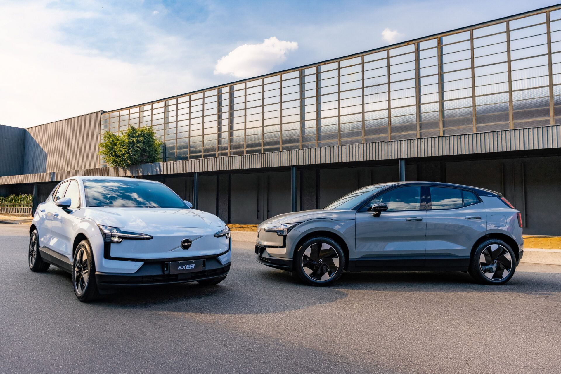 Dois SUVs da Volvo estacionados com uma fachada moderna ao fundo, um branco e um cinza.