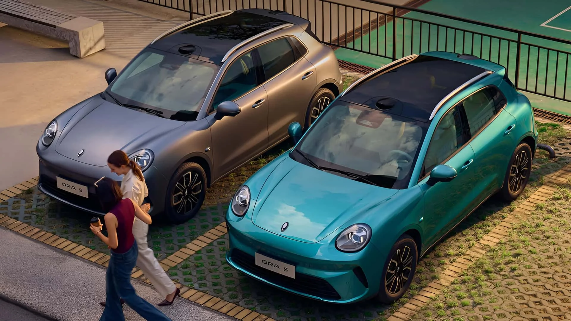 Dois carros, um prateado e um azul, estacionados em um local ao ar livre, com mulheres conversando ao lado.