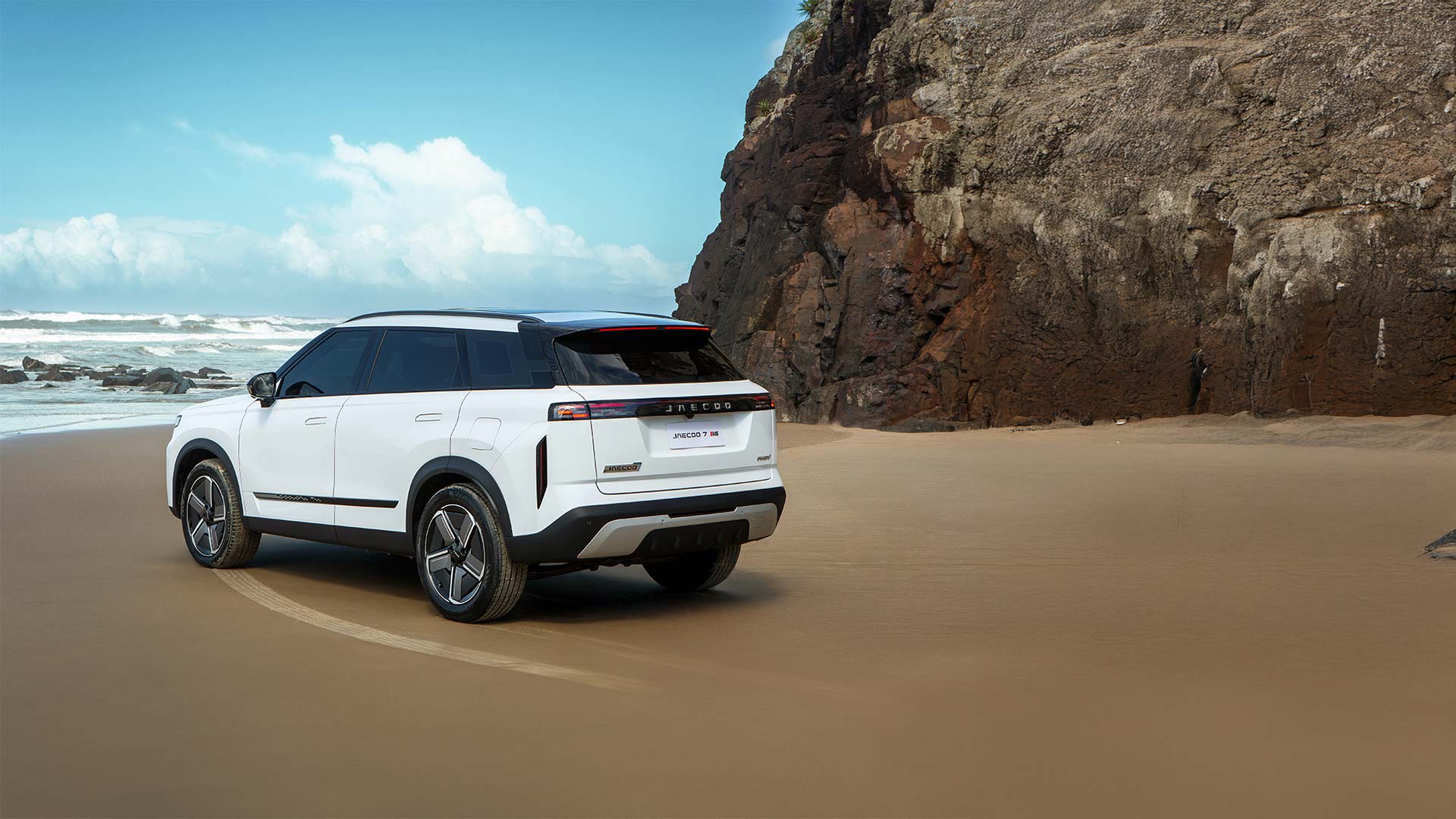 SUV branco estacionado na areia de uma praia, com uma formação rochosa ao fundo e céu azul com nuvens.