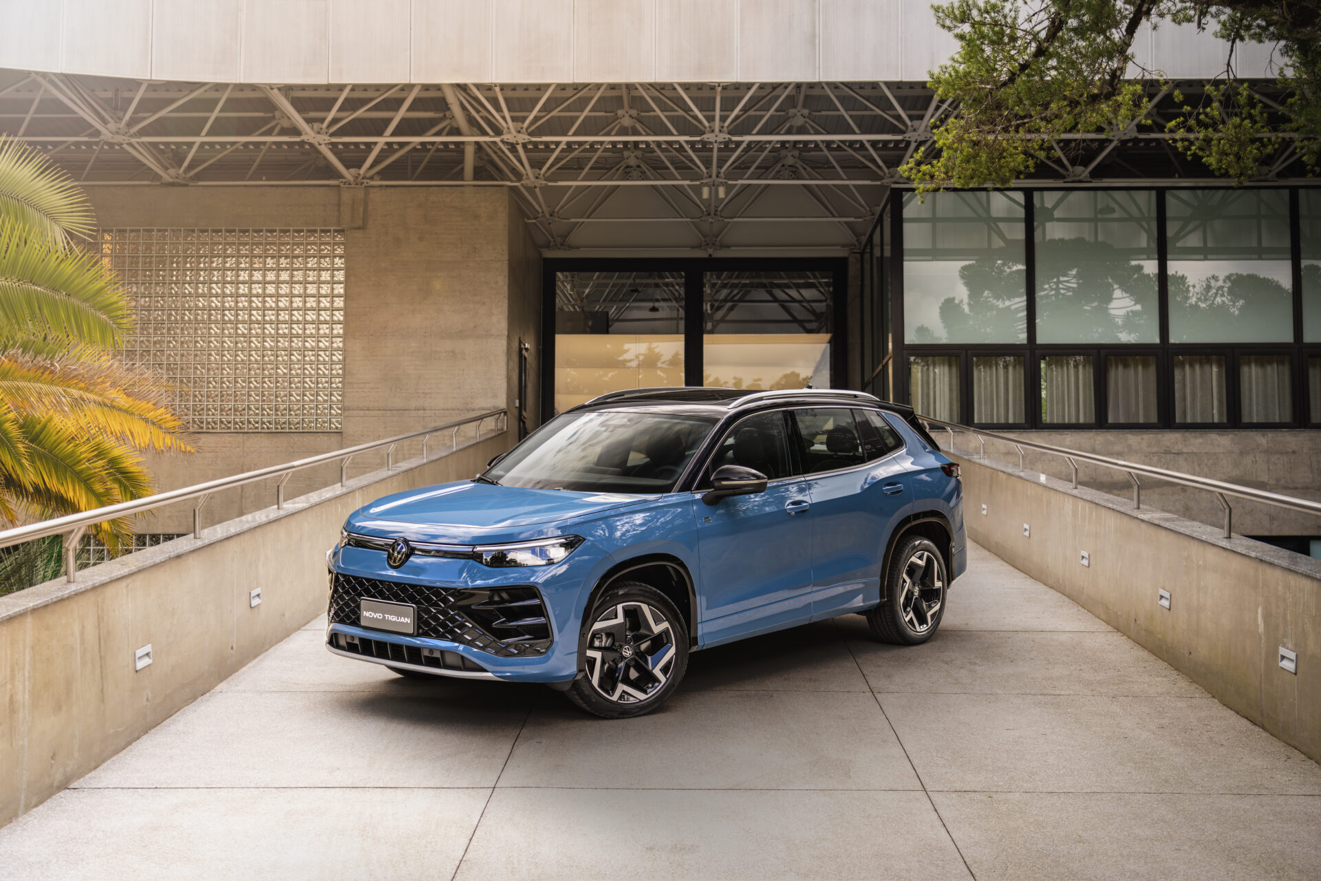 Um carro SUV azul estacionado em uma ampla rampa, com uma estrutura moderna ao fundo e árvores ao lado.