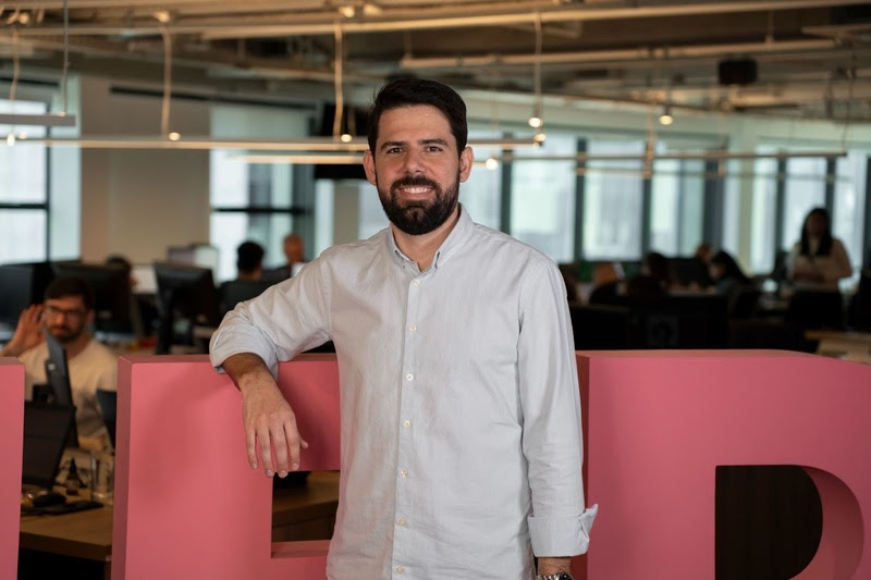 Homem com barba e camisa clara posando em um escritório moderno, com pessoas ao fundo e um grande letreiro rosa.