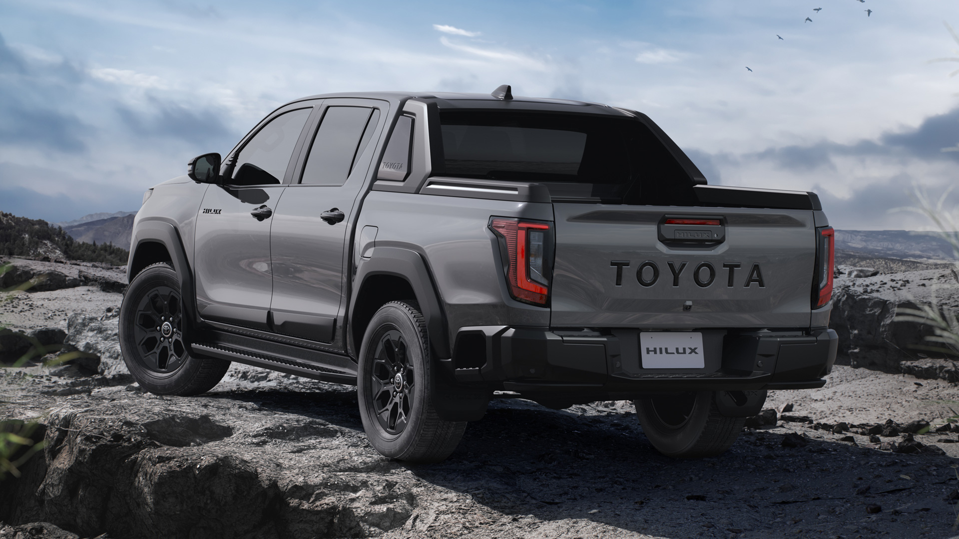 Uma picape Toyota Hilux cinza estacionada em terreno rochoso, mostrando a traseira e lateral do veículo.