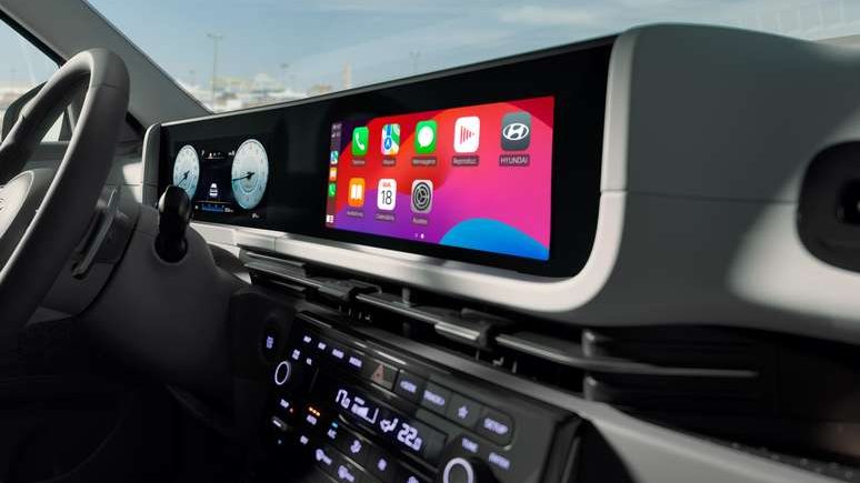 Painel interno de um carro mostrando um display digital com ícones de aplicativos, volante moderno e céu visível pela janela.