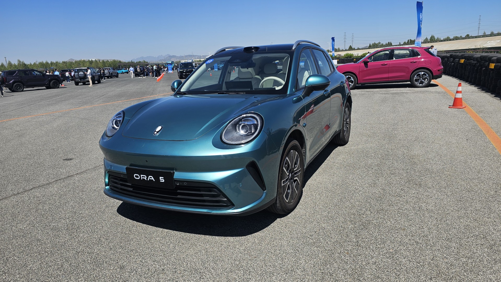 Carro elétrico ORA S em cor verde em exibição em um evento ao ar livre, com outros veículos ao fundo.