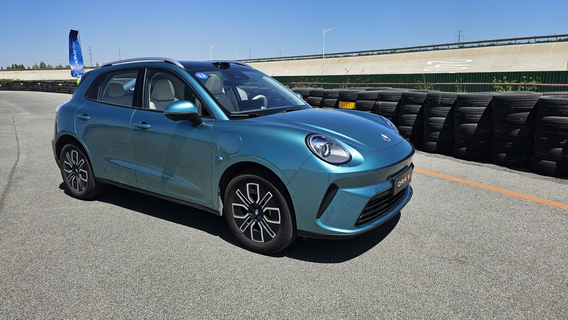 Carro hatchback azul claro estacionado em uma pista de corrida, com pneus empilhados ao fundo e céu limpo.