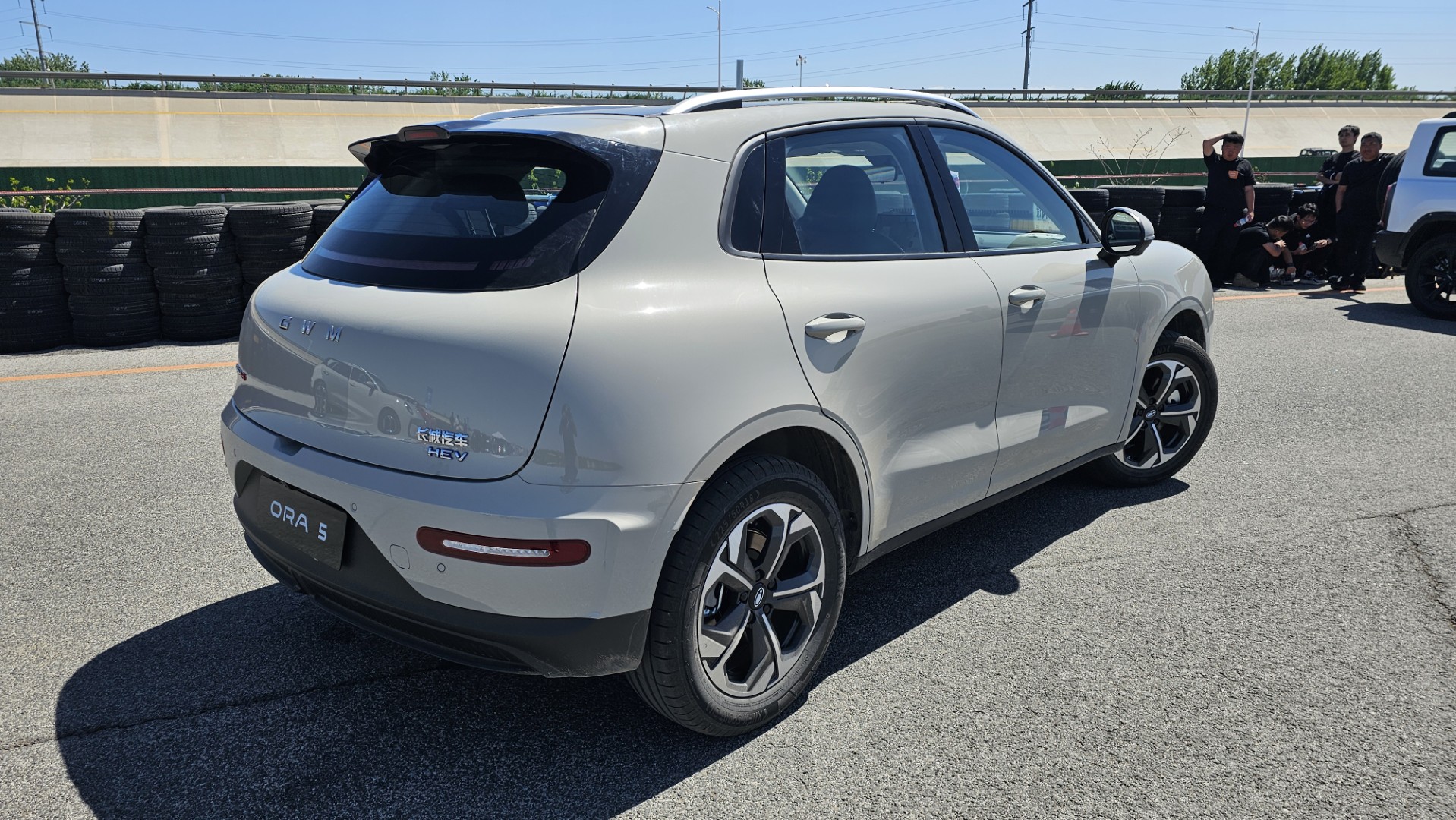 Um carro compacto elétrico na cor prata, visto de lado, com detalhes em preto e rodas esportivas. Ao fundo, há uma pilha de pneus e algumas pessoas vestindo roupas escuras.