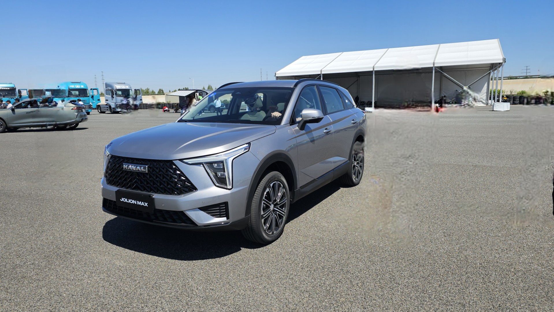 Um SUV prateado Haval Jolion Max estacionado em um evento ao ar livre, com caminhões ao fundo e uma tenda montada.