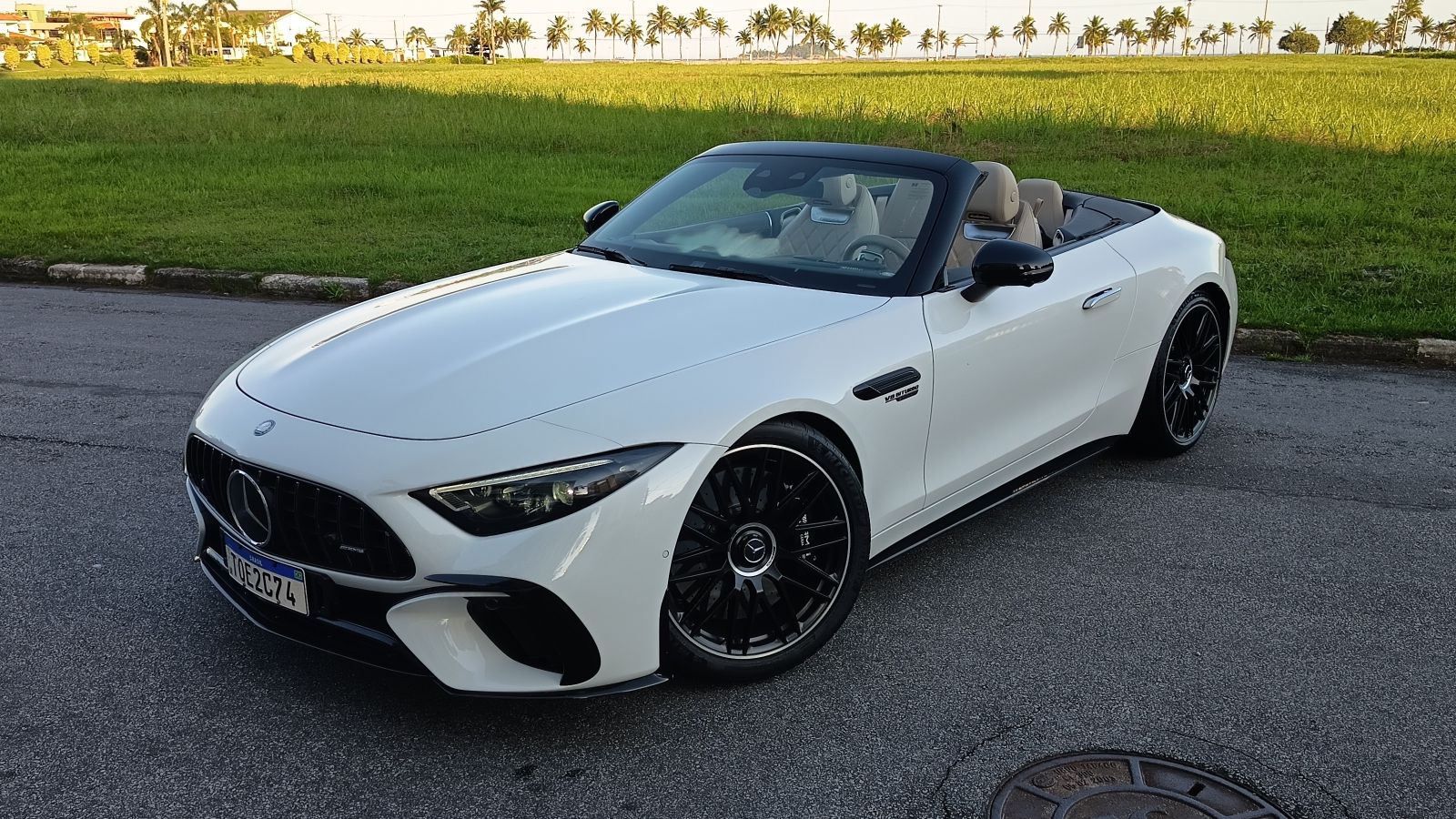 Carro conversível branco da marca Mercedes-Benz, modelo esportivo, estacionado em uma estrada com gramado e palmeiras ao fundo.
