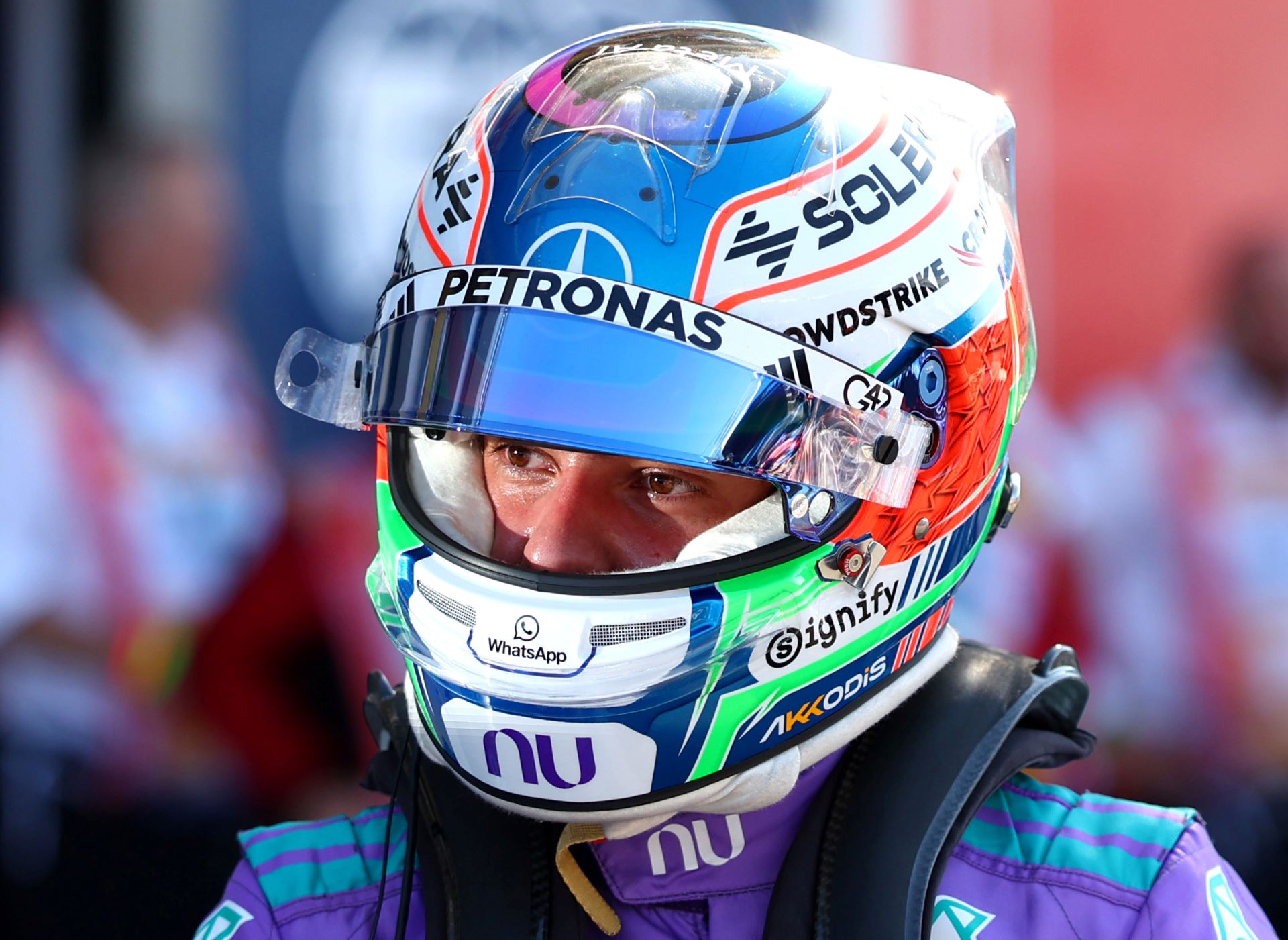 Piloto de automobilismo usando capacete colorido e macacão de corrida, com foco em seu rosto enquanto se prepara para a corrida.