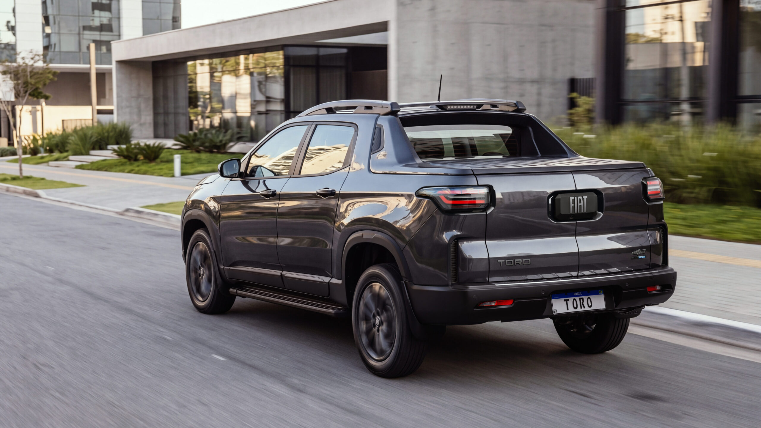 Um carro utilitário esportivo Fiat Toro cinza se deslocando na rua, com edifícios modernos ao fundo.