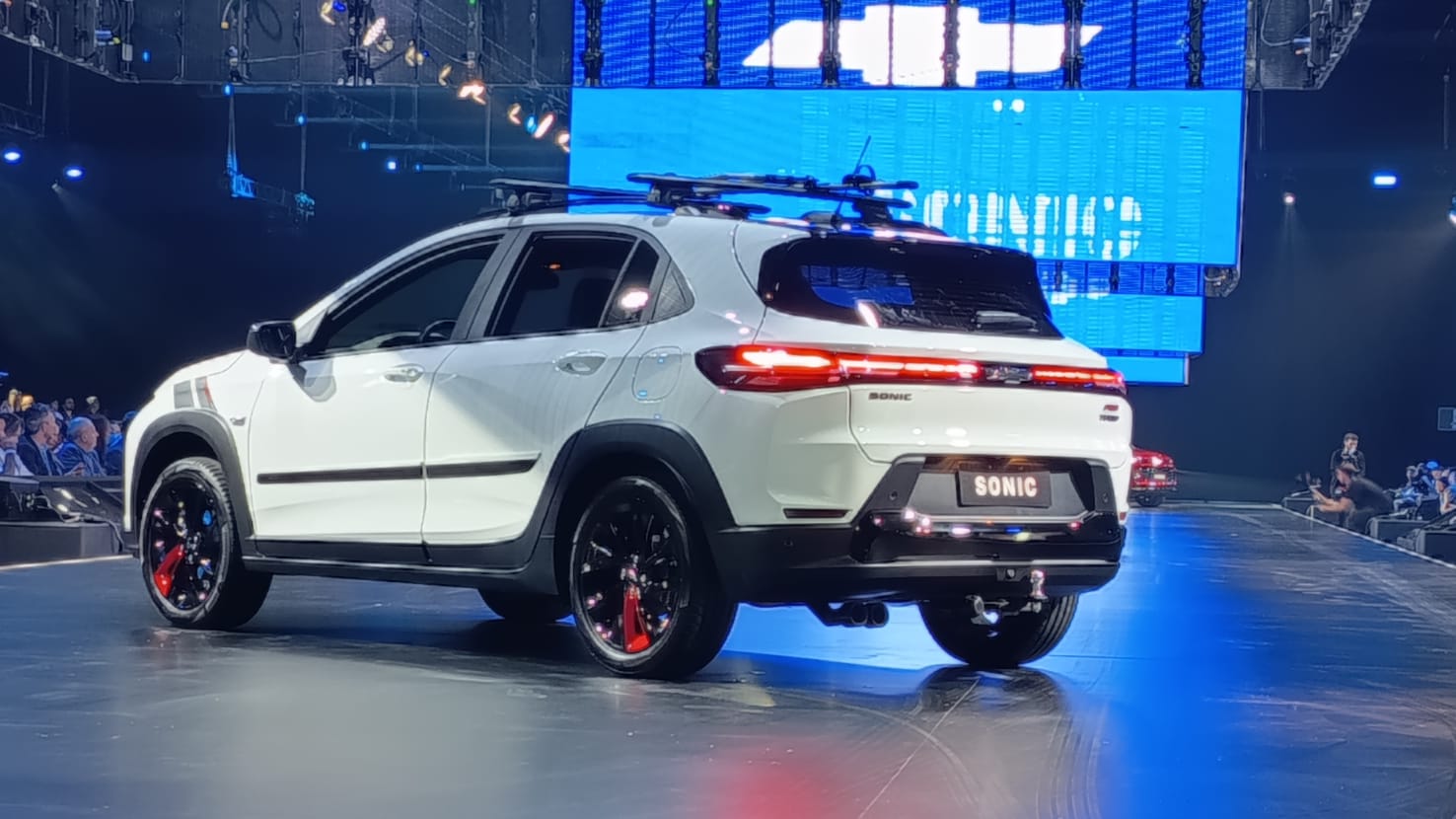 Um carro SUV branco com detalhes esportivos, exibido em um palco iluminado durante um evento, com rodas pretas e um rack de teto.