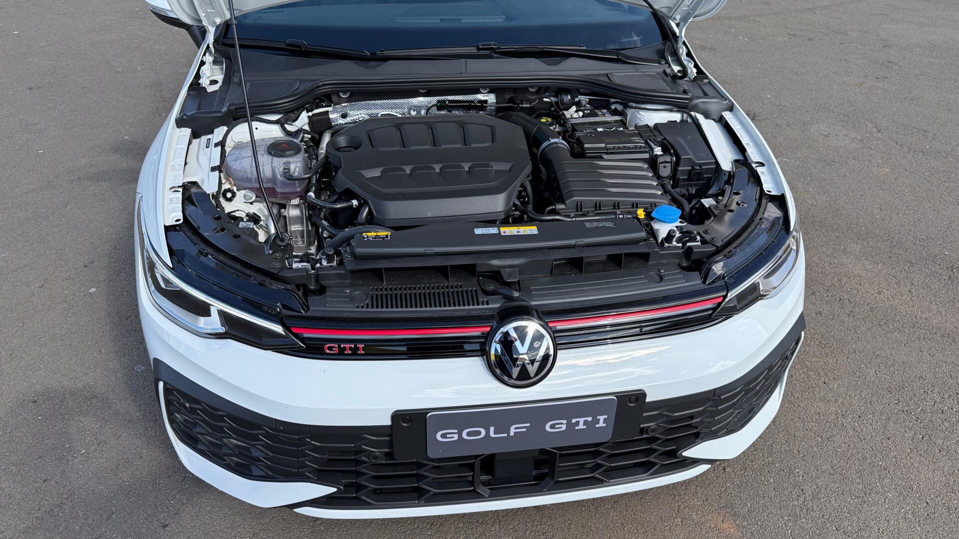 Visualização do motor de um Volkswagen Golf GTI, com capô aberto, mostrando componentes e logo frontal.