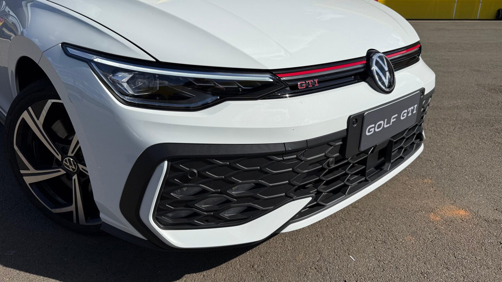 Close-up da frente de um carro Volkswagen Golf GTI branco, destacando o farol, a grade frontal e o emblema GTI.