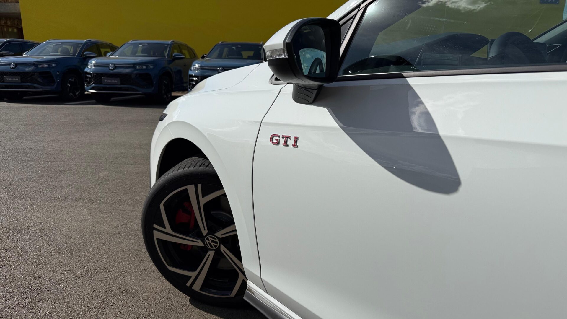 Close-up de um carro branco com o emblema GTI, ao fundo uma fila de SUVs Volkswagen em uma área de estacionamento.