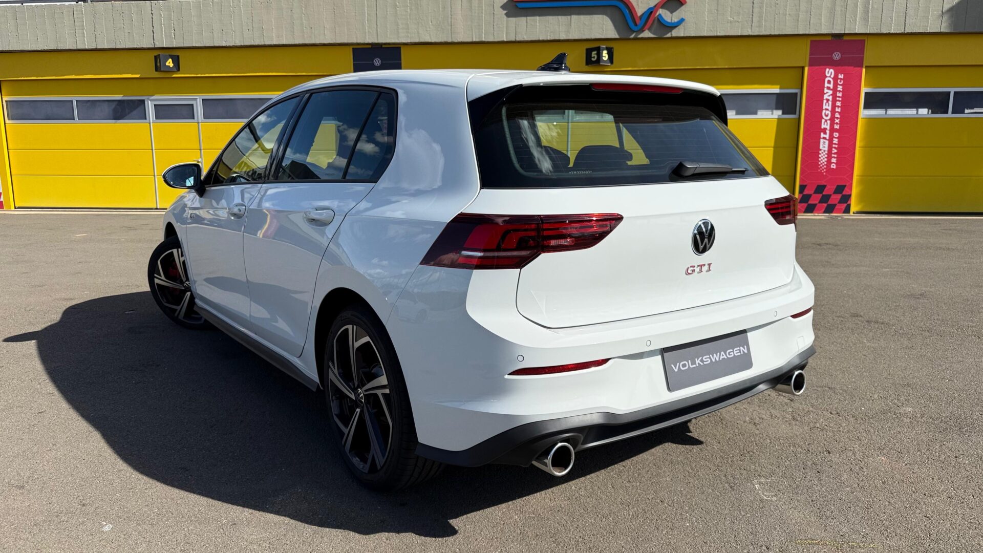 Vista traseira de um carro Volkswagen GTI branco estacionado em um local com fundo amarelo.
