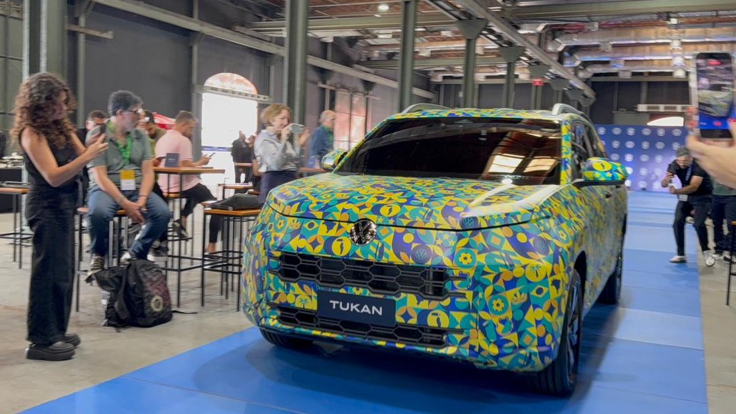 Um carro modelo Tukan colorido com padrões vibrantes em uma sala de eventos, rodeado por pessoas tirando fotos.