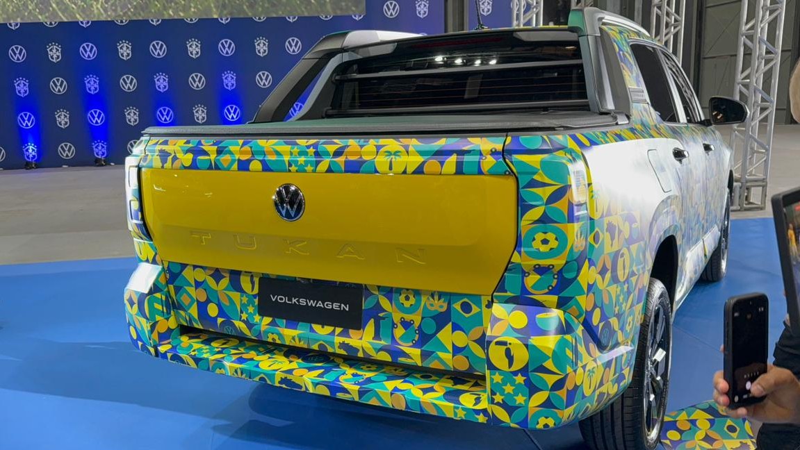 Parte traseira de uma caminhonete Volkswagen Tukan decorada com padrões coloridos, exibida em um evento com fundo iluminado em azul.