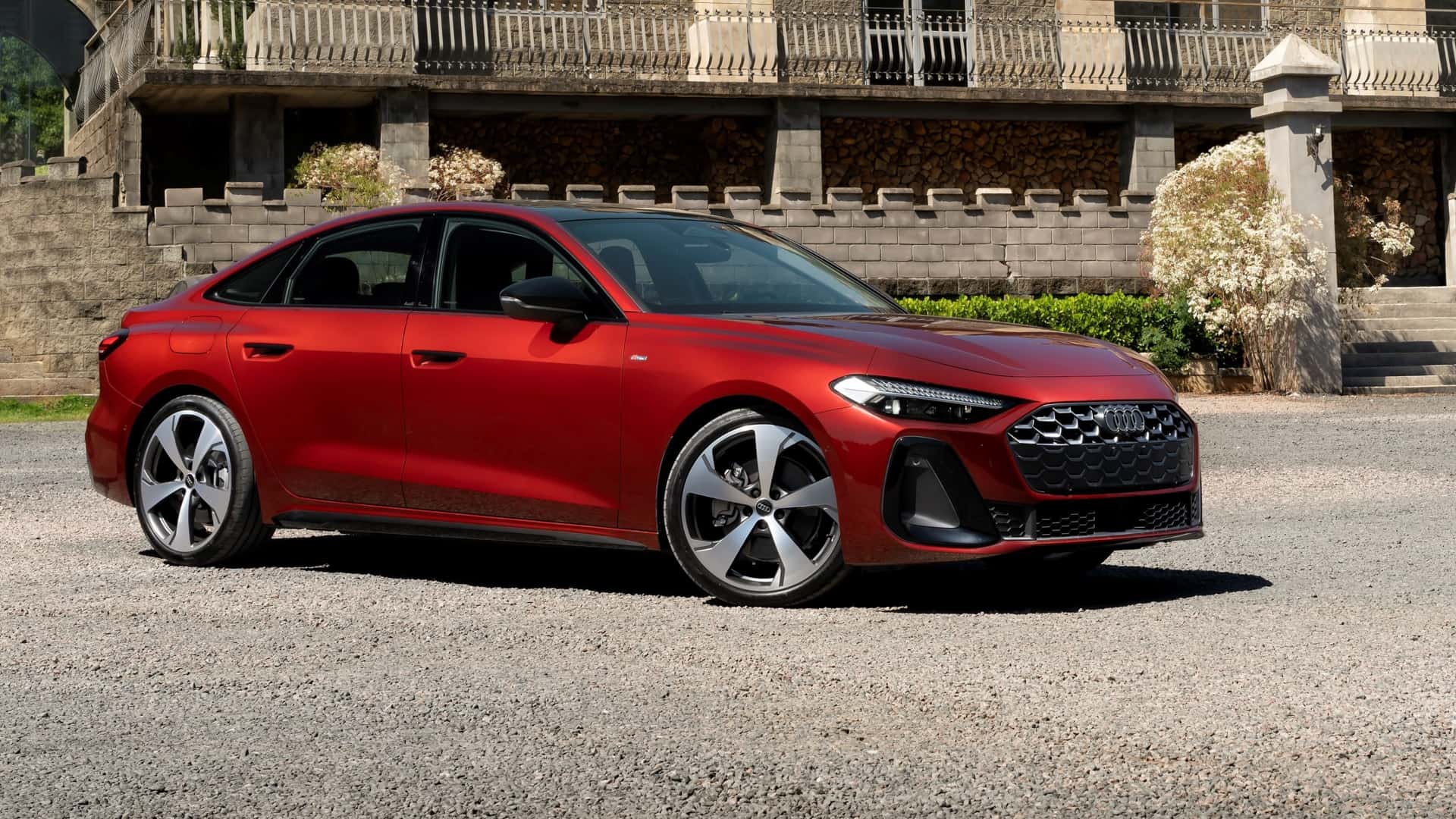 Carro sedan vermelho estacionado em uma área de cascalho, com uma construção antiga ao fundo.
