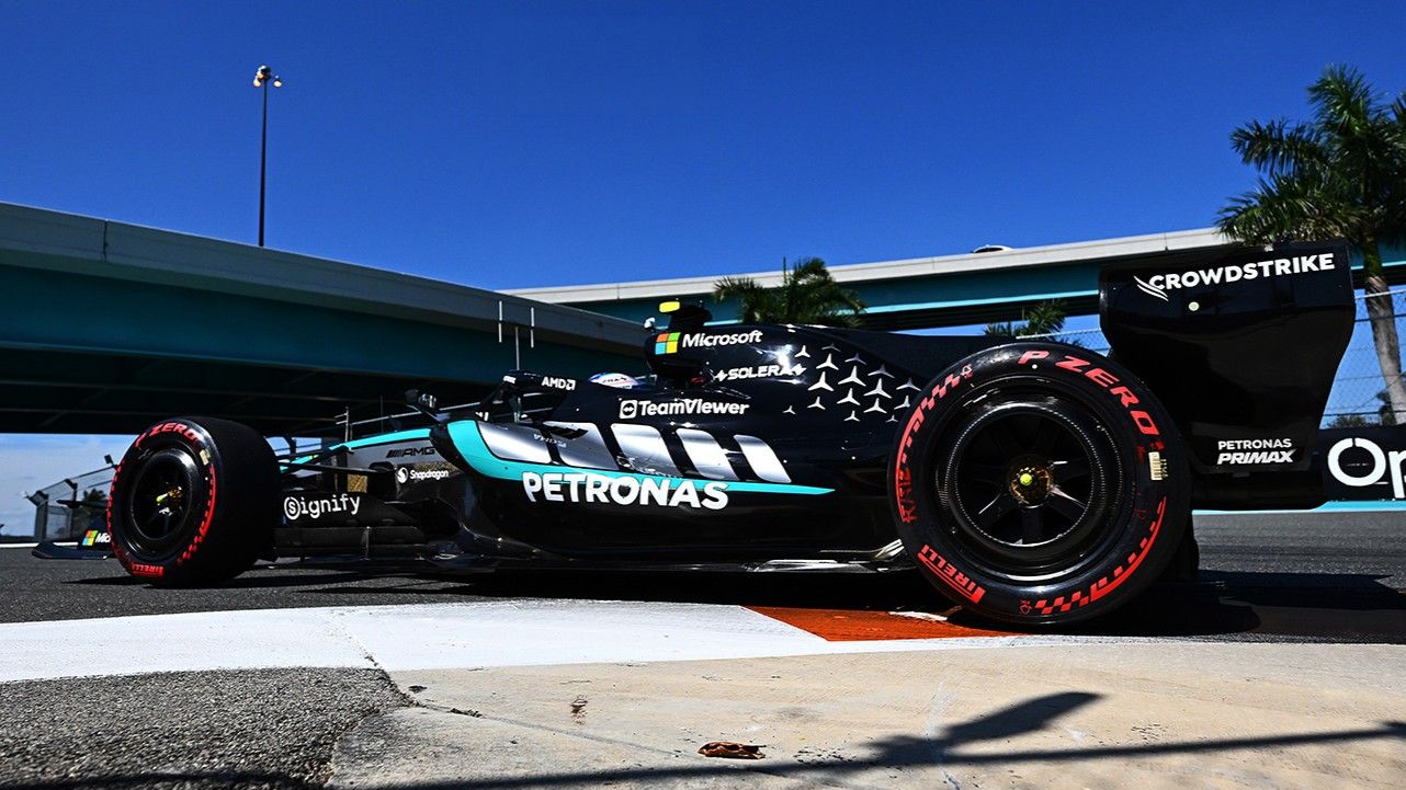 Carro de Fórmula 1 da equipe Mercedes com patrocínios visíveis, na pista sob um céu azul ensolarado.