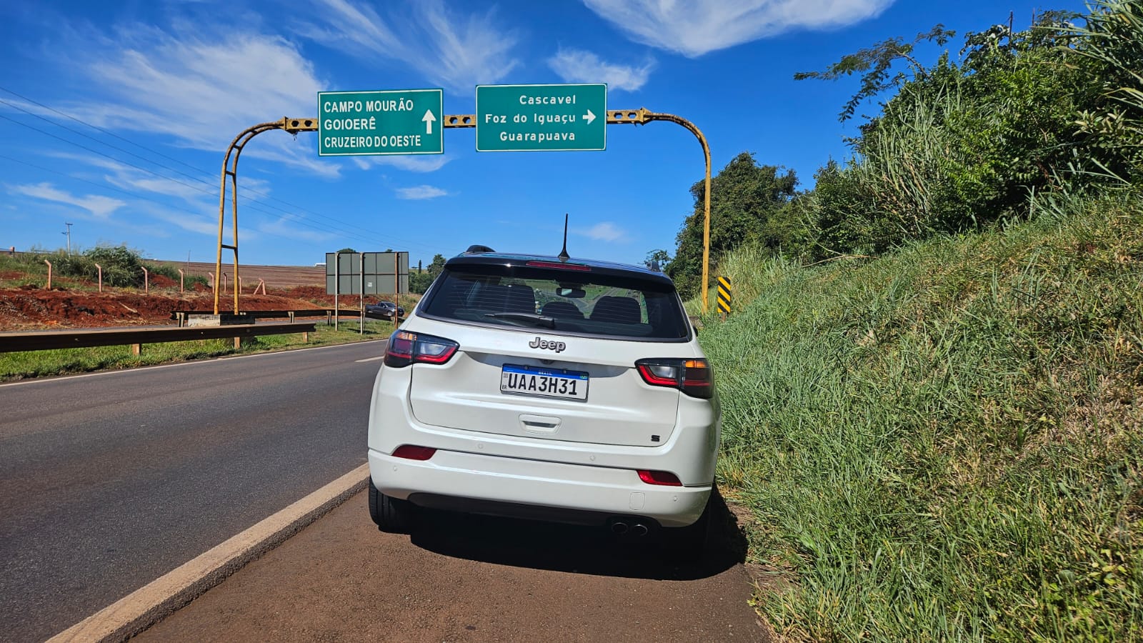 Foto de um carro Jeep estacionado à beira da estrada, com placas de sinalização para as cidades Campo Mourão, Goioerê, Cruzeiro do Oeste, Cascavel, Foz do Iguaçu e Guarapuava ao fundo, sob um céu azul.