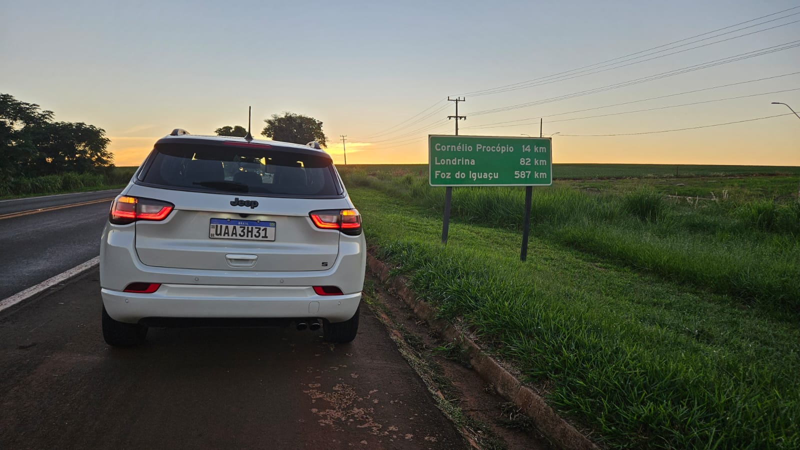 Um carro branco estacionado ao lado de uma estrada, com um painel de sinalização indicando distâncias para Cornélio Procópio, Londrina e Foz do Iguaçu ao fundo.