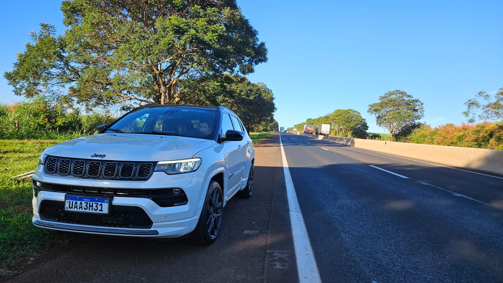 Um carro SUV branco modelo Jeep estacionado ao lado de uma estrada em um dia ensolarado, com árvores verdes ao fundo.