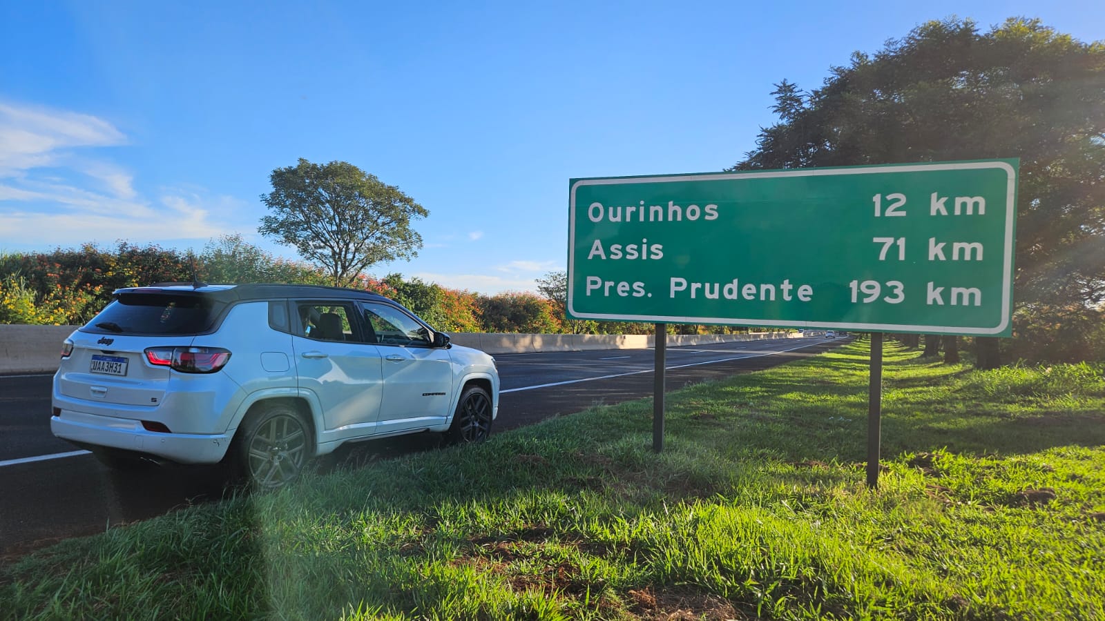 Um carro branco ao lado de uma placa de estrada indicando distâncias para Ourinhos (12 km), Assis (71 km) e Presidente Prudente (193 km).