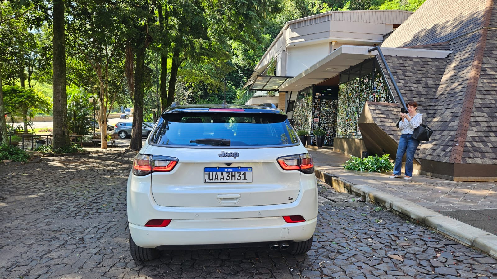 Um carro Jeep branco estacionado em uma área arborizada, com uma mulher em pé ao lado, segurando um celular. Ao fundo, uma fachada de loja com muitas adesivos.