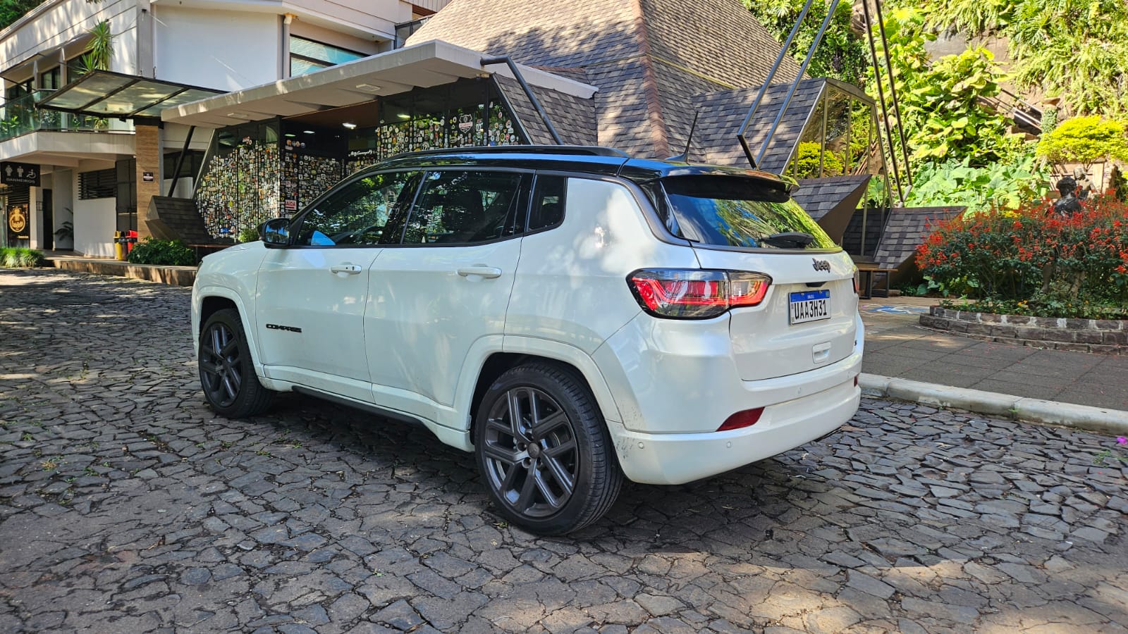 SUV branca estacionada em uma área com pedras, ao lado de um edifício moderno e jardins.