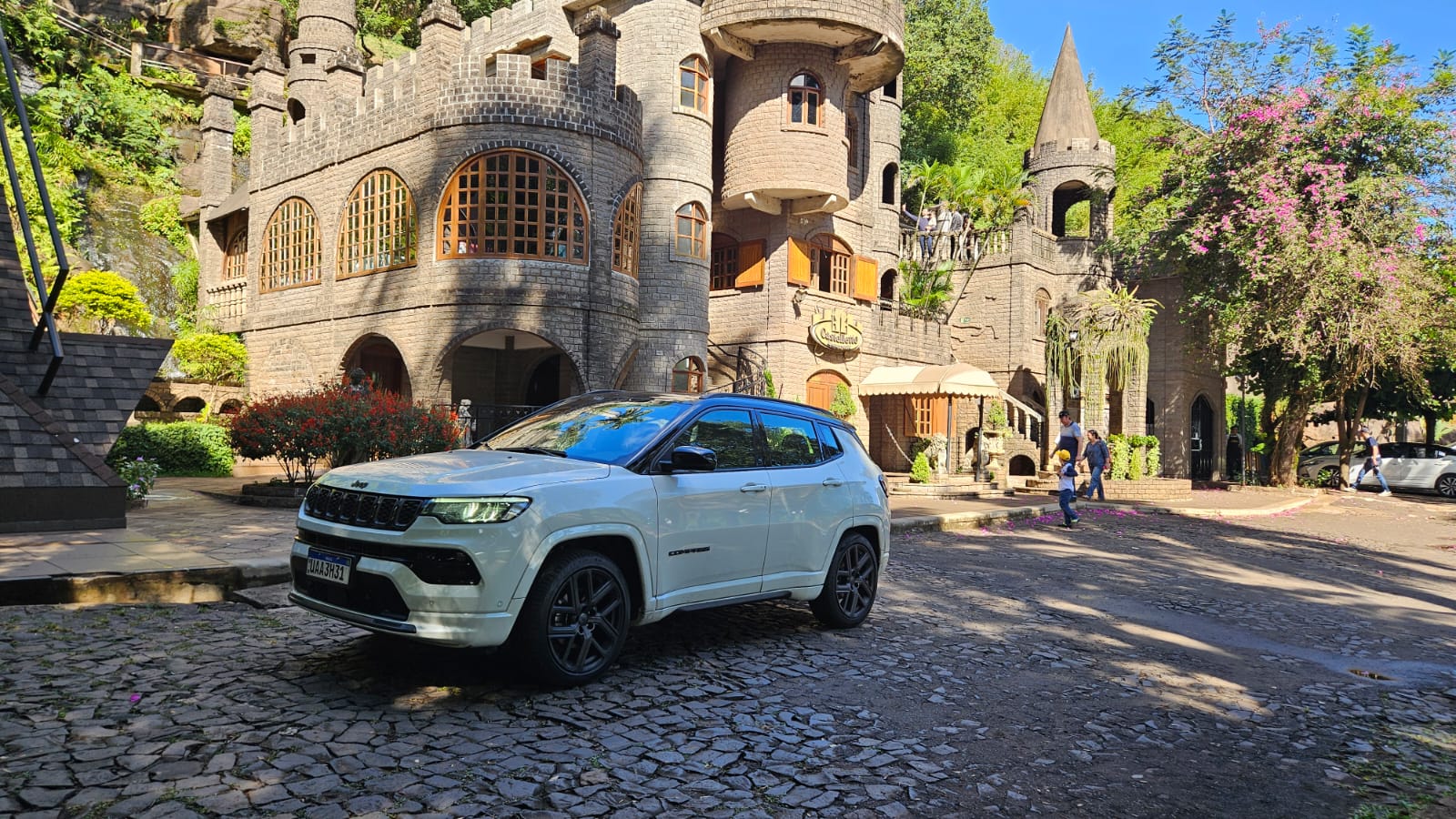 Um carro SUV branco estacionado em frente a um edifício em estilo de castelo, cercado por vegetação e flores.