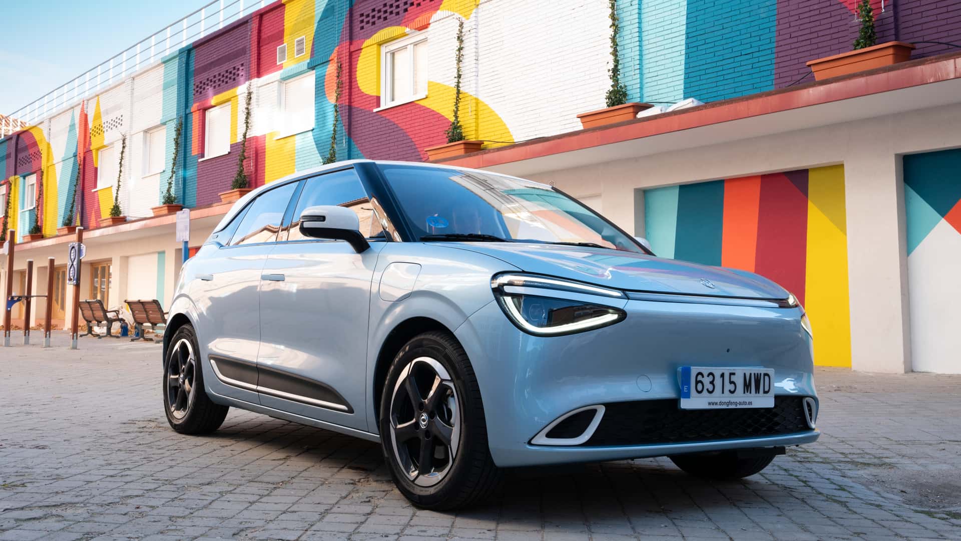 Um carro compacto azul claro estacionado em uma rua com murais coloridos ao fundo.