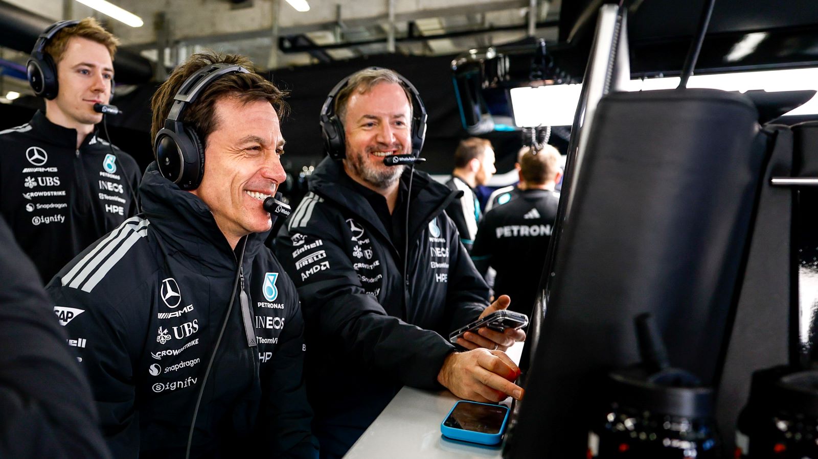 Imagem de membros da equipe Mercedes durante um evento de automobilismo, sorrindo e usando fones de ouvido enquanto observam um monitor.
