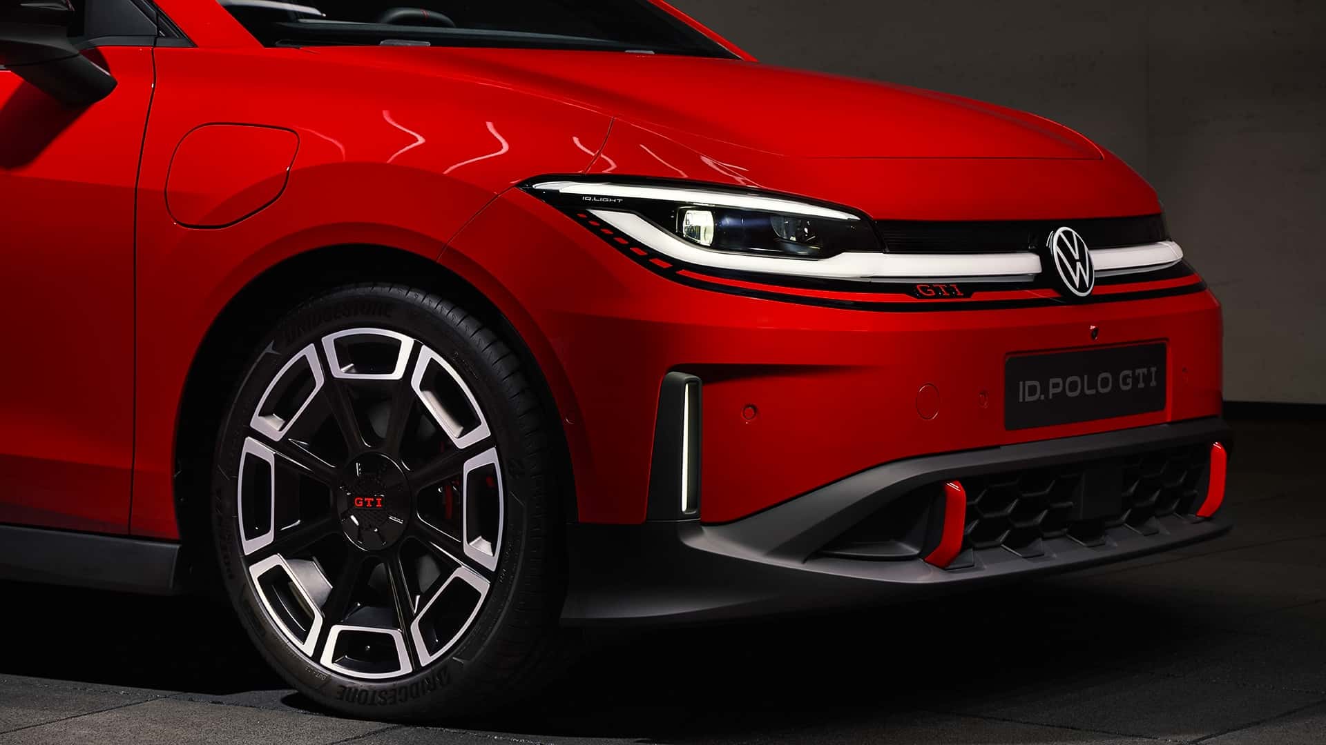 Close-up do Volkswagen ID. Polo GTI vermelho, destacando a roda com detalhes em preto e cromado, faróis com design moderno e logo da Volkswagen na grade frontal.