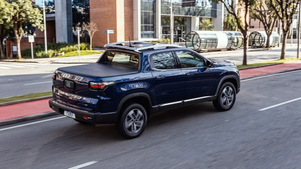 Uma picape Fiat Toro azul em movimento em uma rua urbana, com edifícios ao fundo.