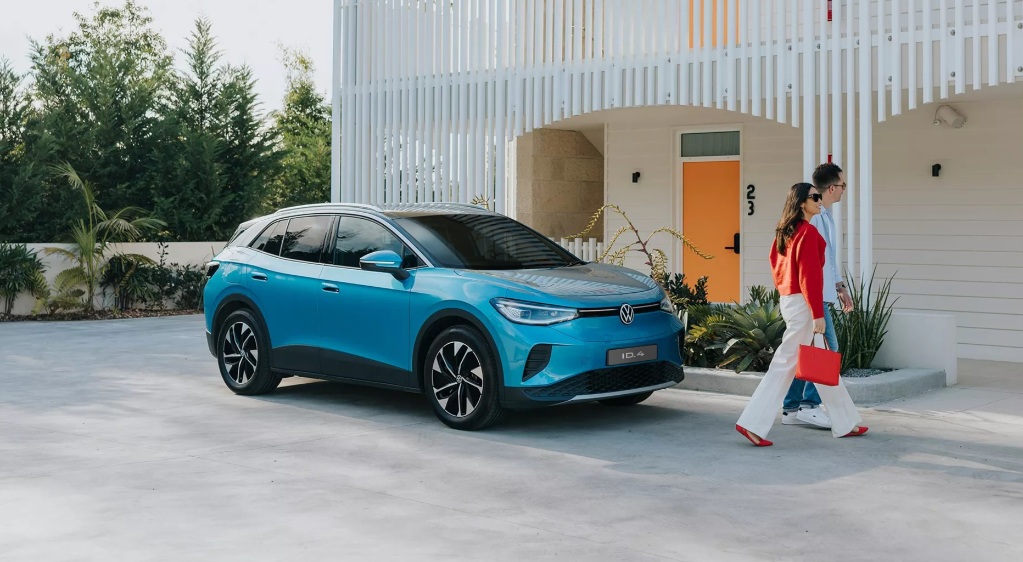 A blue electric SUV parked in a driveway with two people walking by, surrounded by greenery and modern architecture.