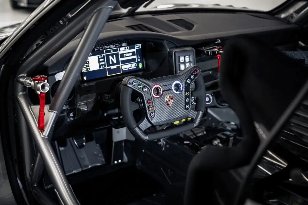 Close-up view of a racing car's cockpit featuring a carbon fiber steering wheel with multiple buttons, a digital display dashboard, and safety features like a roll cage.