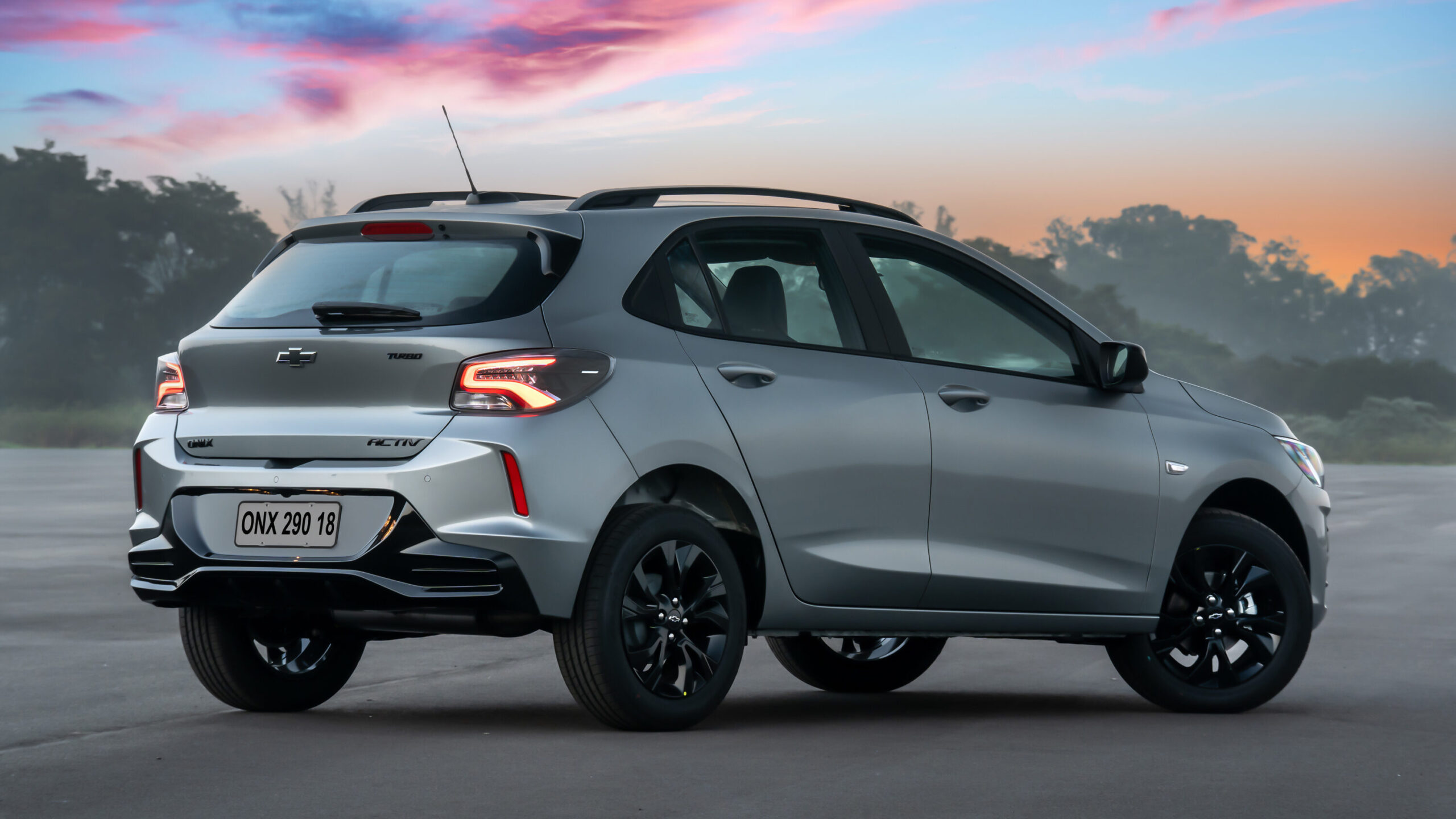A silver Chevrolet car, model Onix Activ, parked on pavement with a scenic sunset background featuring pink and orange hues.