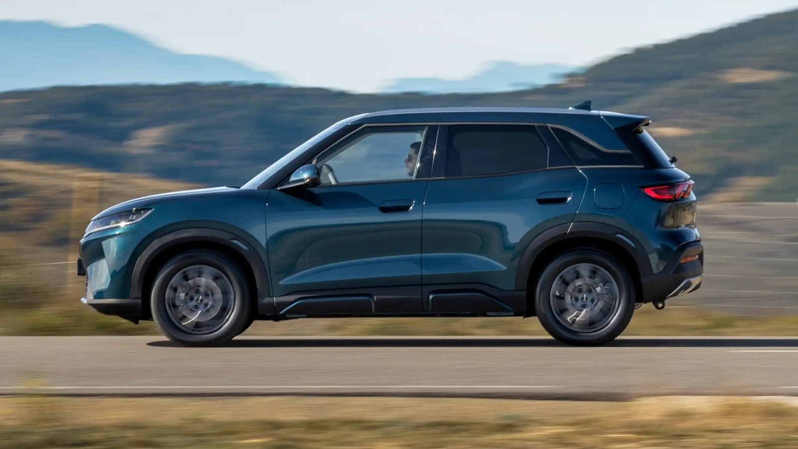 Side view of a teal SUV driving on a scenic road with mountains in the background.