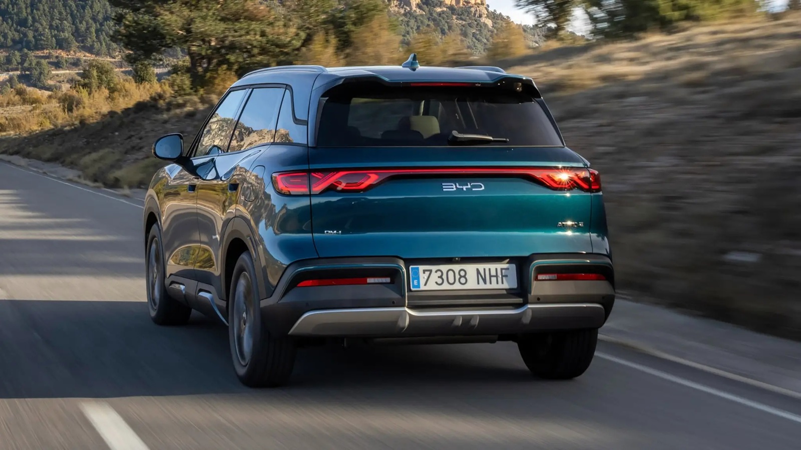 A teal-colored BYD vehicle driving along a winding road surrounded by nature, showcasing its rear design with distinctive taillights and license plate.