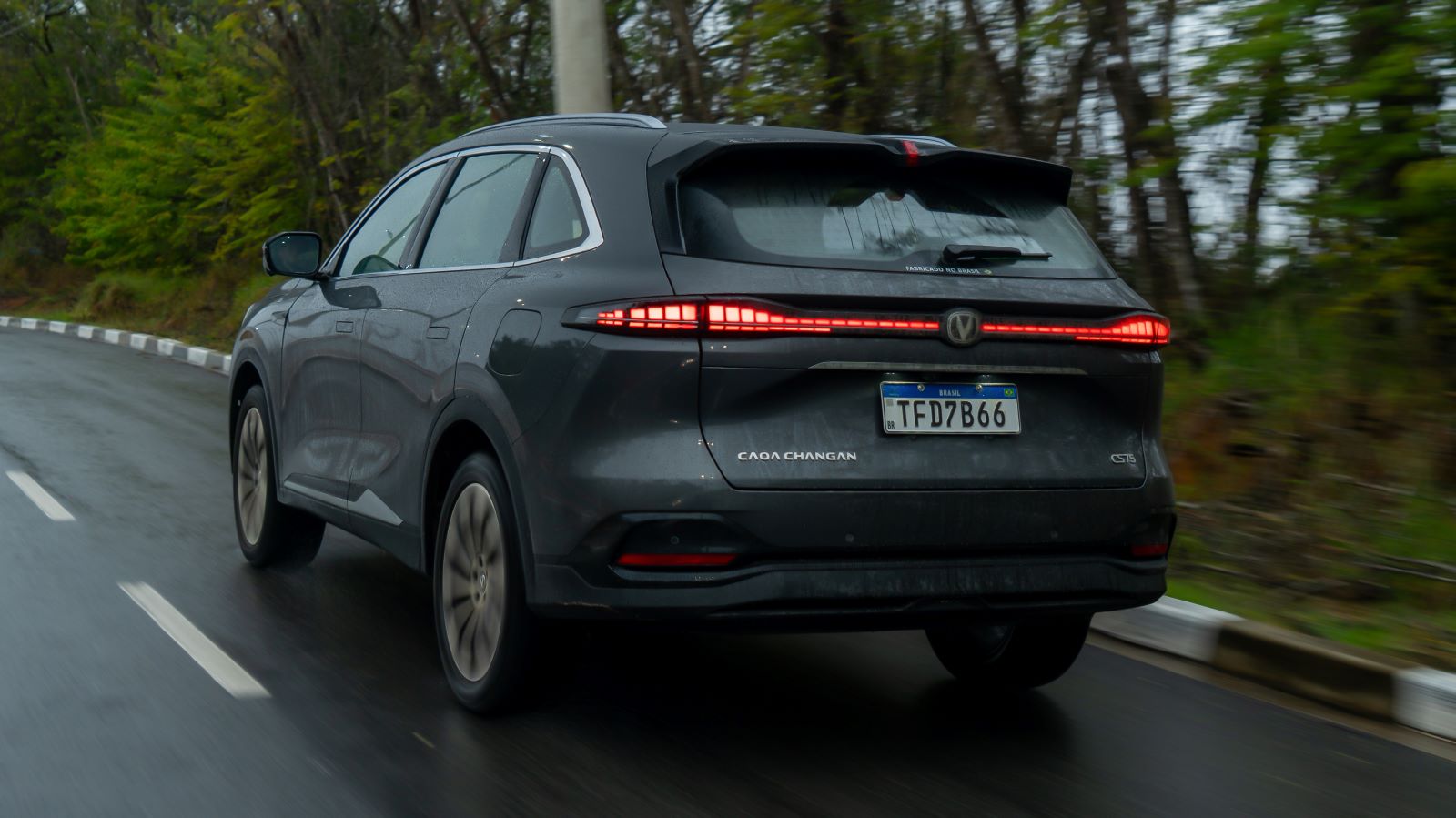 Um carro SUV cinza, com as luzes traseiras acesas, em uma estrada curva, cercado por árvores verdes, em um dia chuvoso.