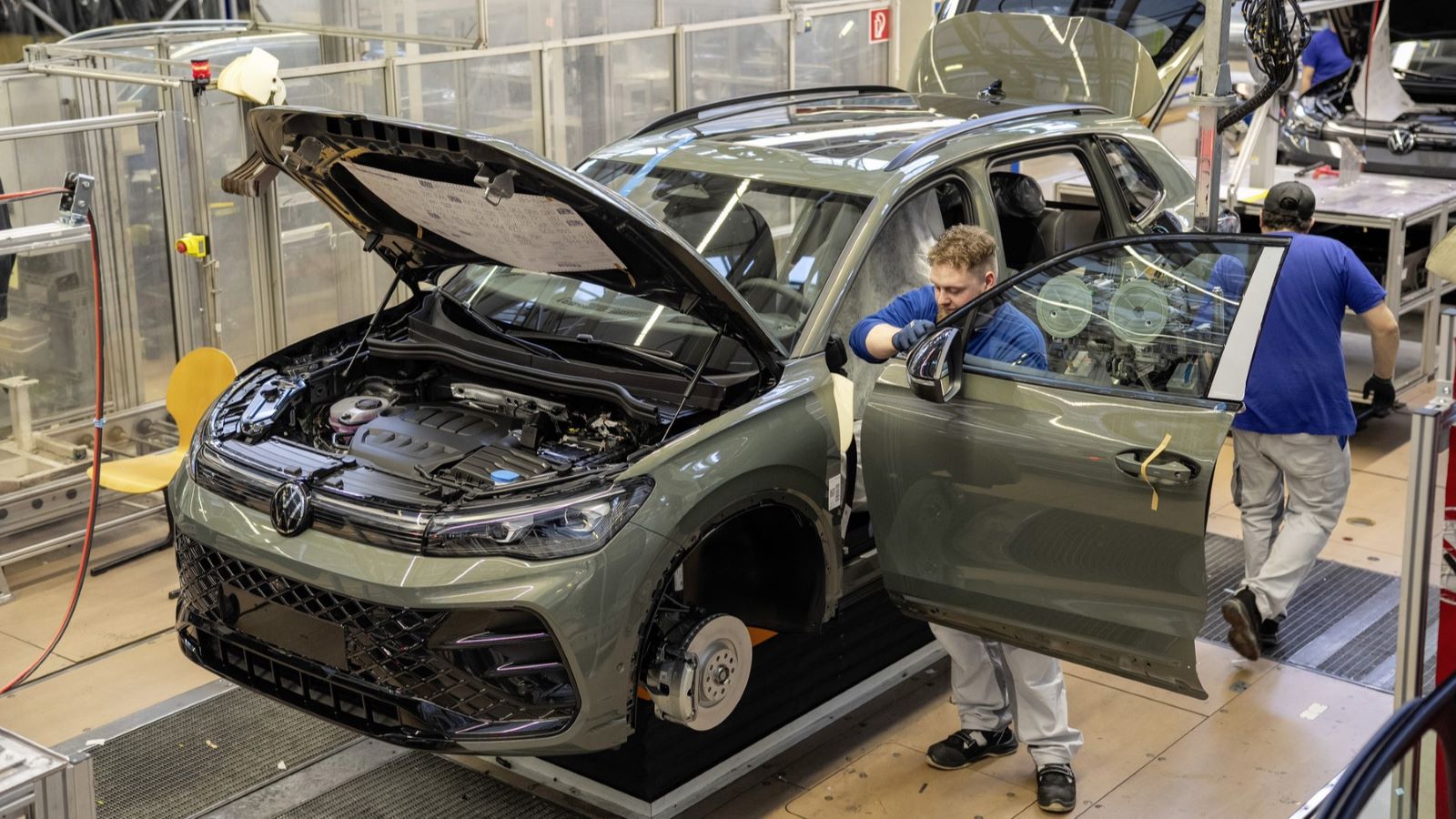 Foto de uma linha de montagem de automóveis, mostrando um SUV com o capô aberto e trabalhadores realizando ajustes.