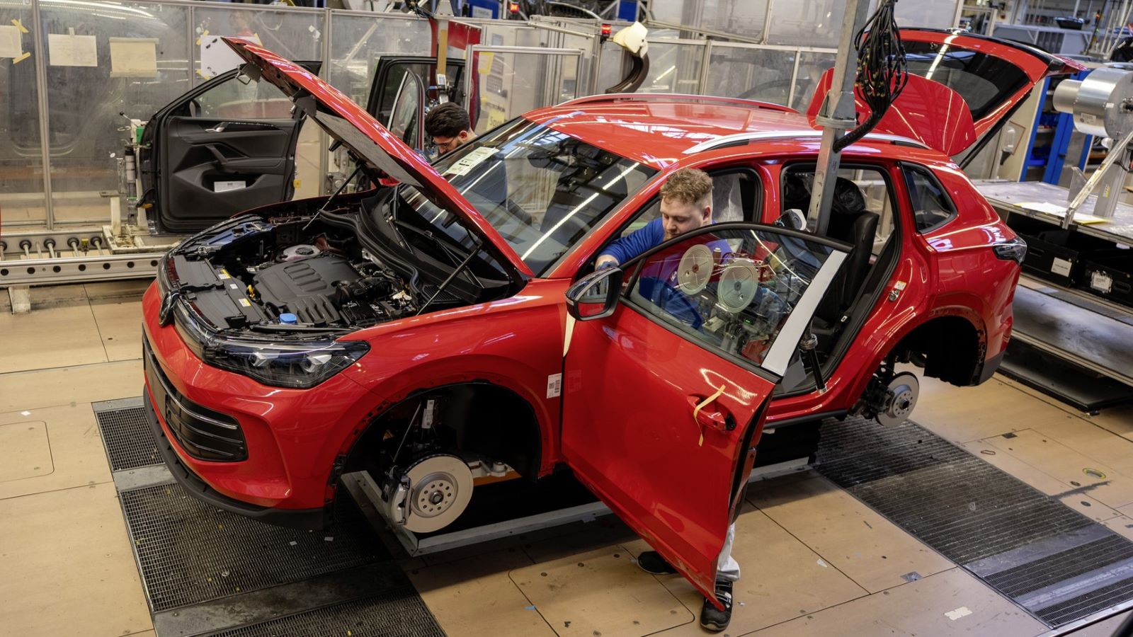 Um carro vermelho sendo montado em uma linha de produção, com dois trabalhadores realizando ajustes sob o capô e na porta dianteira.