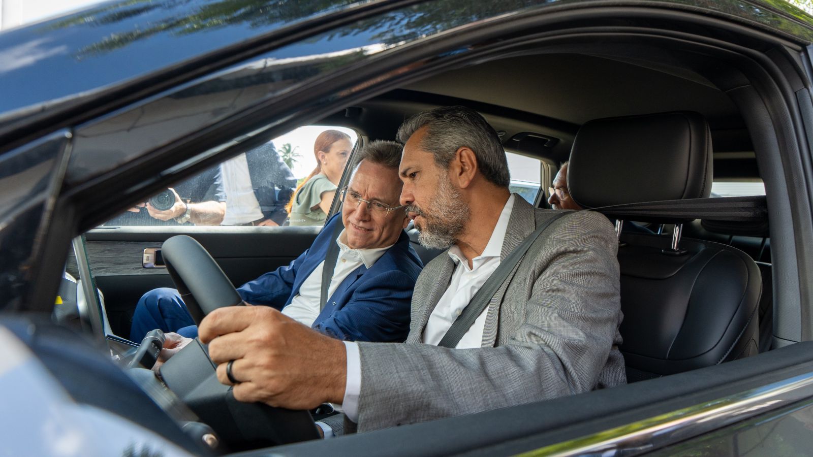  Dois homens sentados no banco da frente de um carro conversando, um deles segurando o volante. 