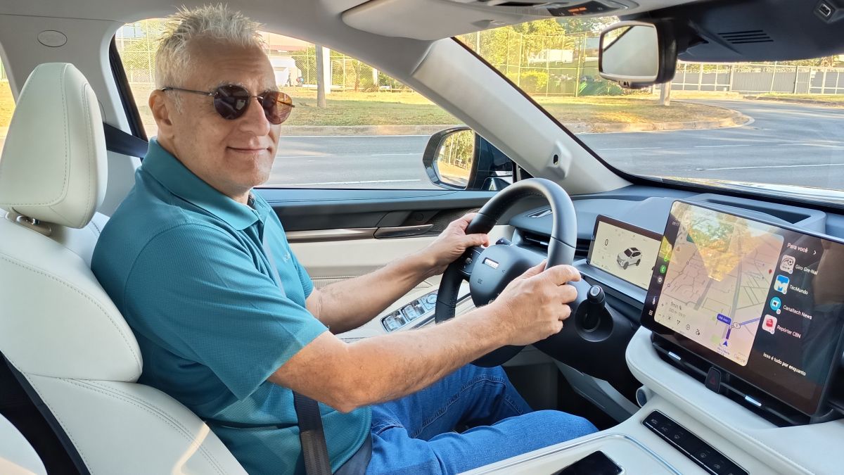 Homem sorridente dirigindo um carro moderno, usando óculos de sol e uma camisa polo azul, com telas digitais visíveis no painel do veículo.