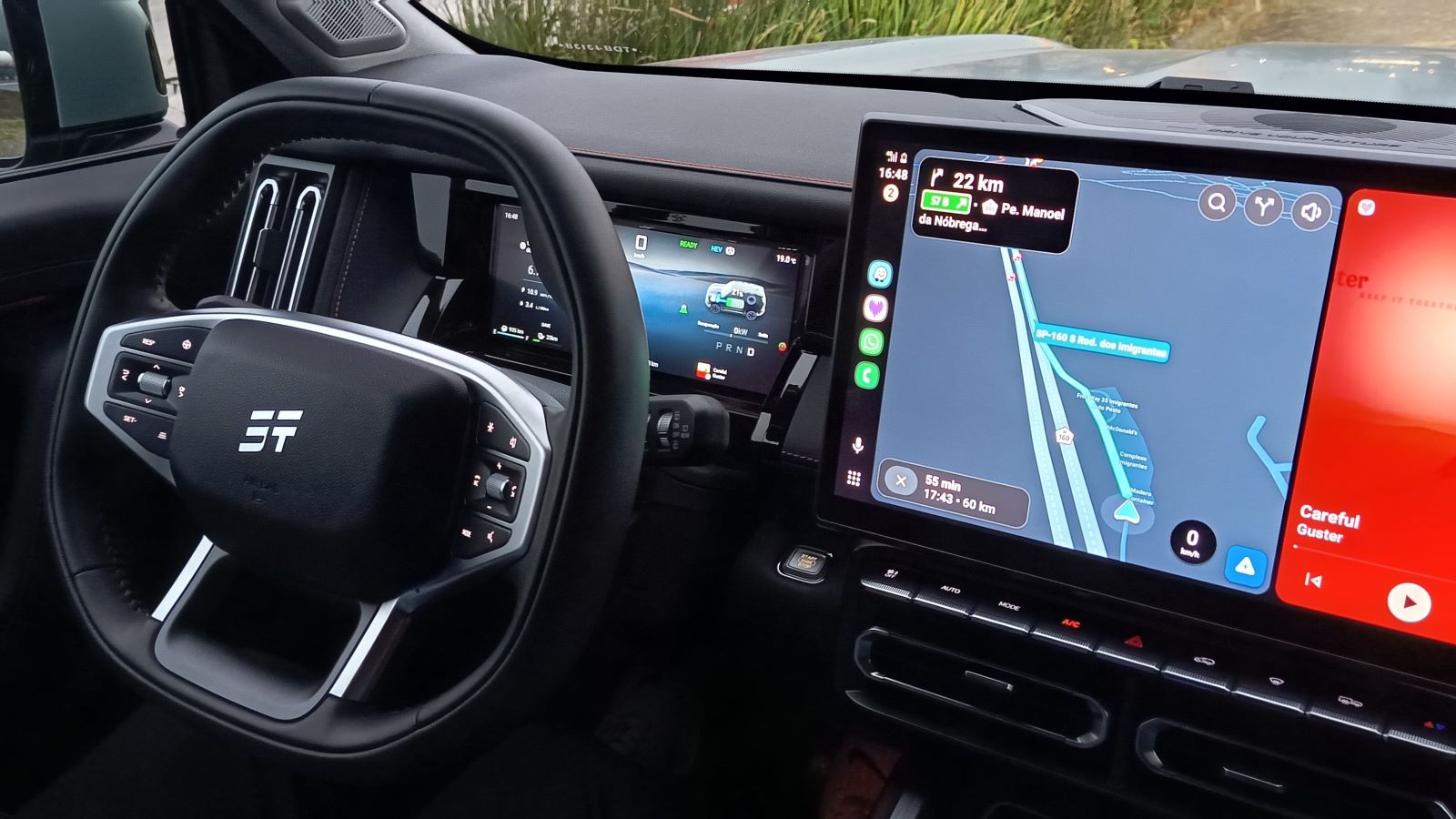 Vista interna de um carro com volante moderno e telas digitais no painel, exibindo informações de navegação e desempenho.