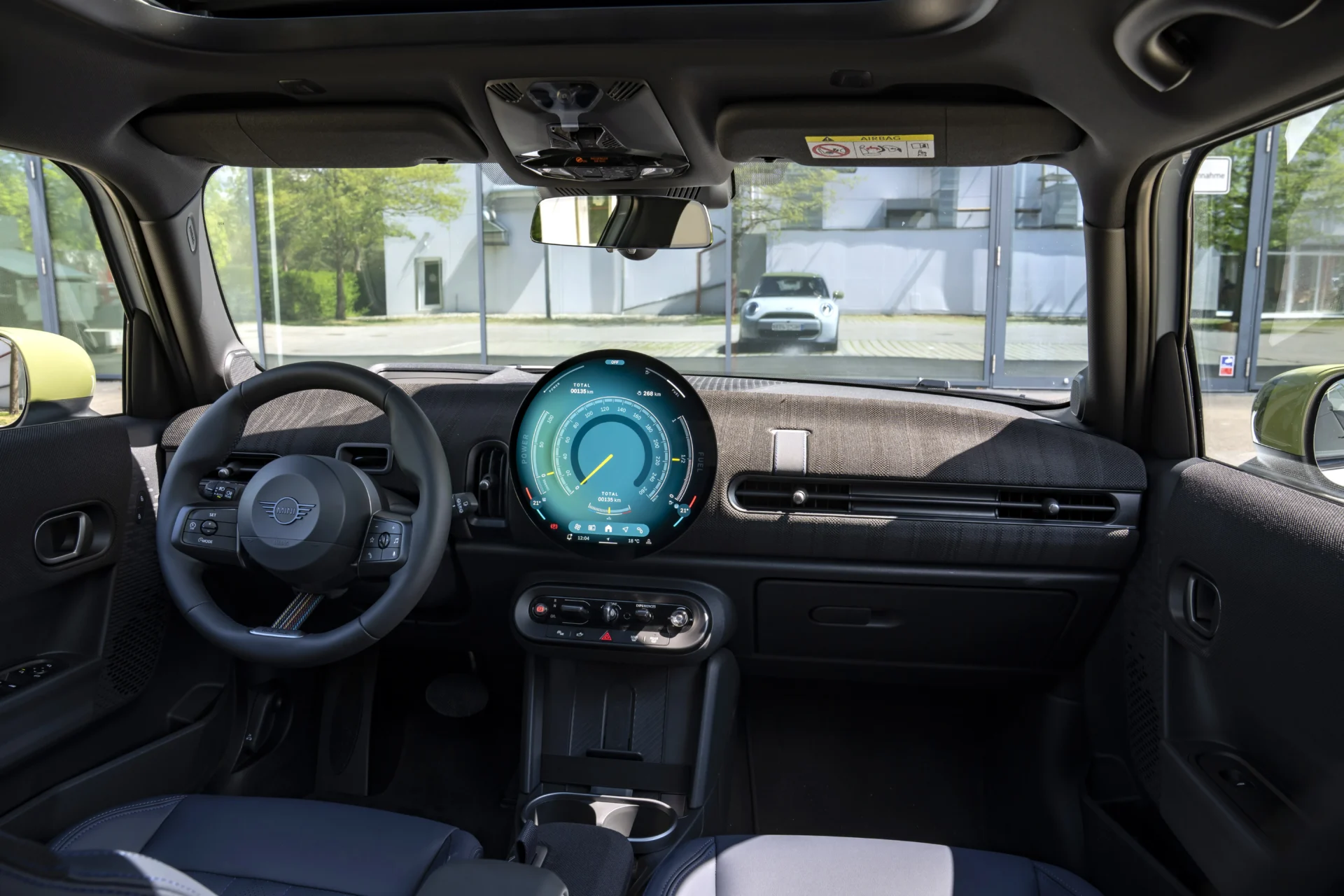 Interior view of a modern car featuring a circular digital dashboard display, steering wheel, and controls. Large windows provide a view of the outside environment.