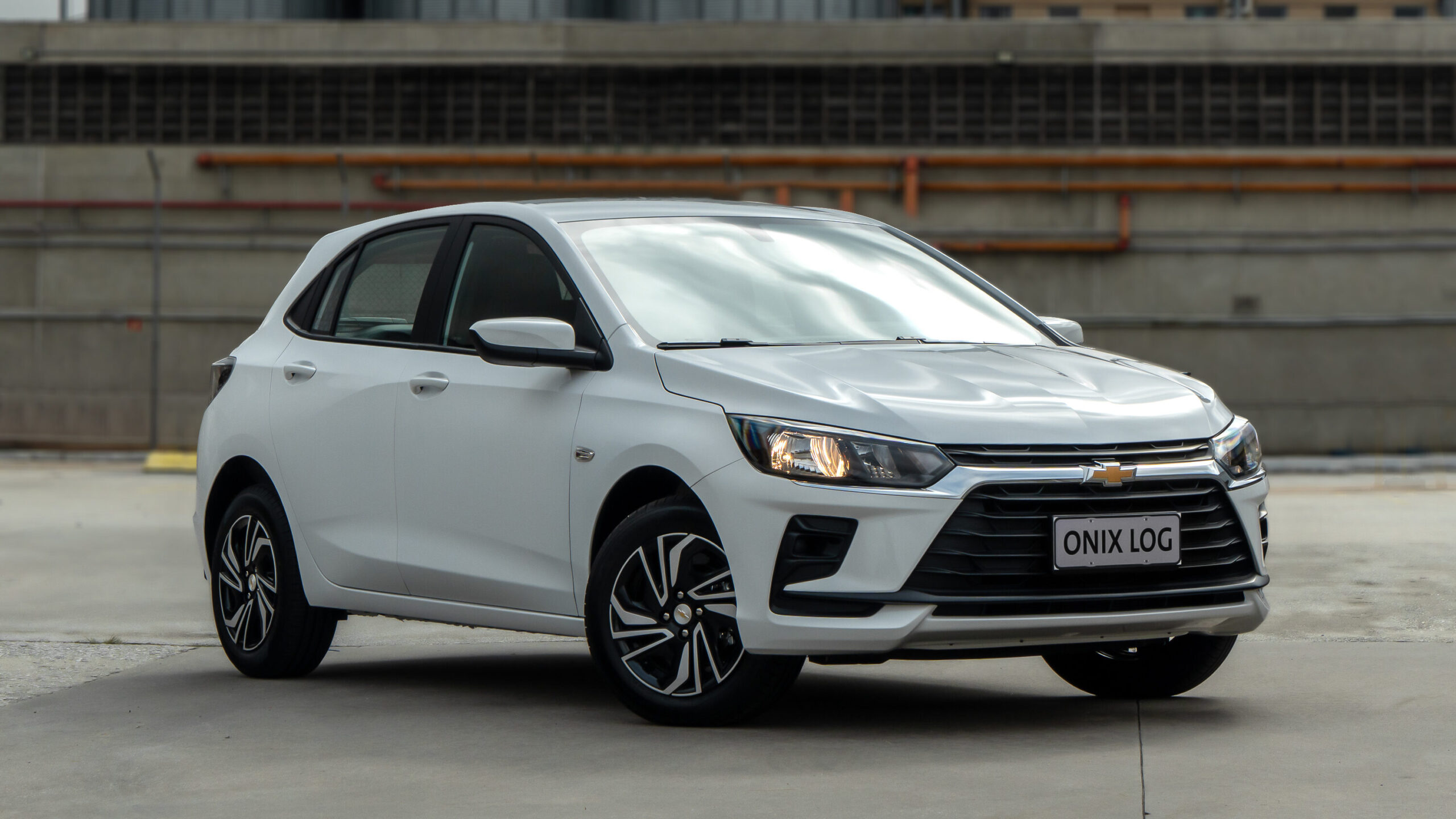 A silver Chevrolet Onix hatchback parked in an industrial area with a modern building in the background.