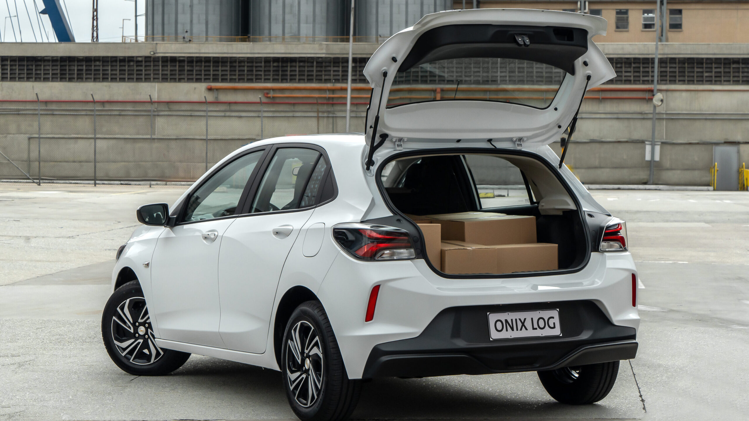A white hatchback car parked with its rear hatch open, revealing cardboard boxes inside the trunk in an industrial setting.