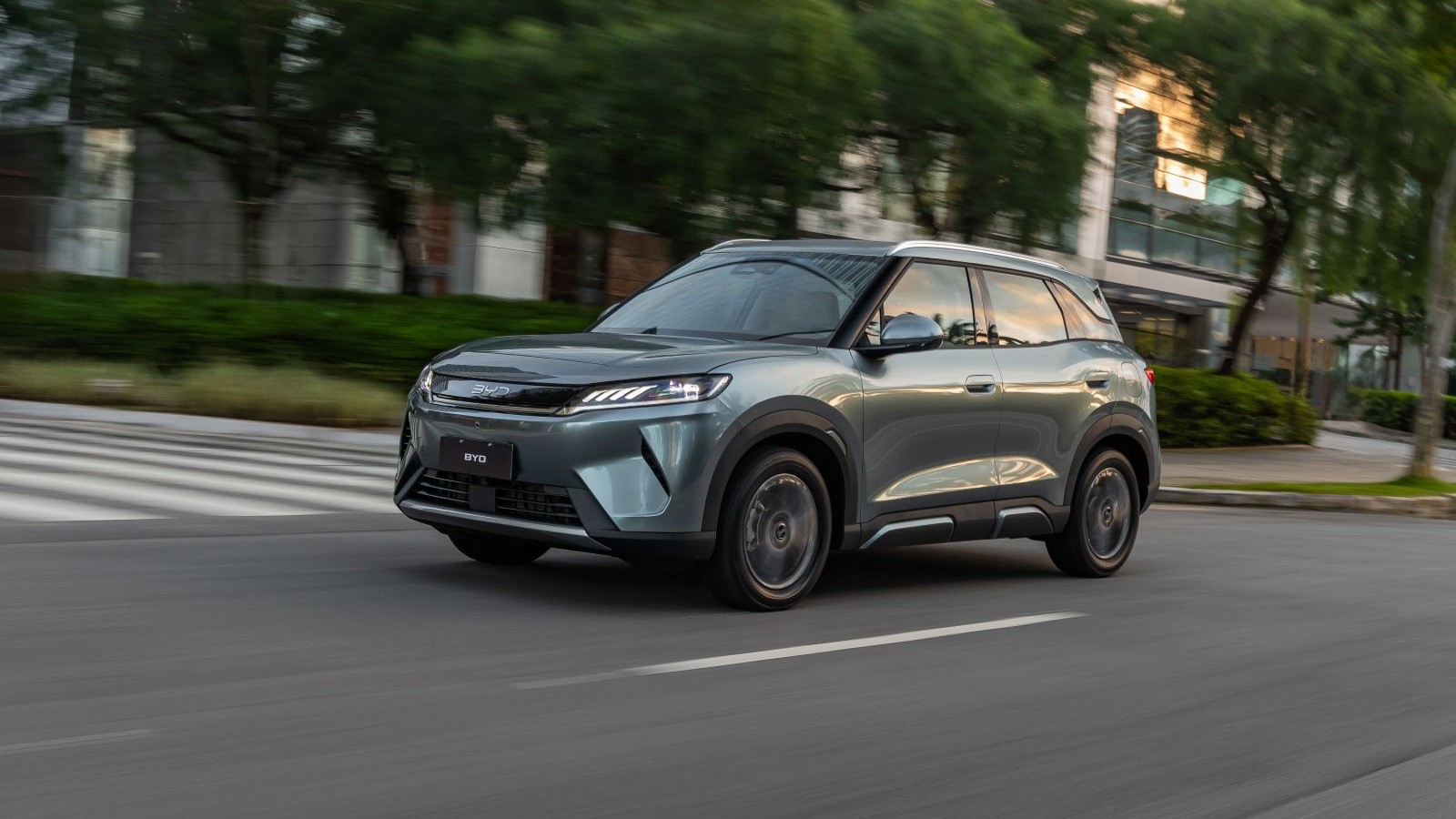Carro SUV cinza em movimento em uma rua urbana, com vegetação e prédios ao fundo.